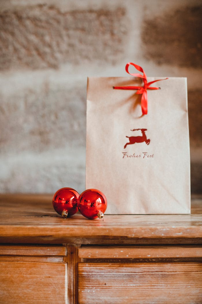 Brown paper gift bag with red ribbon and reindeer print, placed alongside glossy red baubles on a rustic wooden table.
